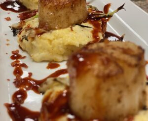 A white plate with cheese grits topped with seared scallops, with a drizzle of BBQ sauce.