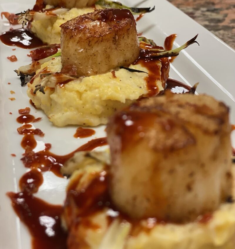A white plate with cheese grits topped with seared scallops, with a drizzle of BBQ sauce.