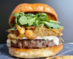 Photo of a French Onion Soup Burger with a bun, burger, cheese, caramelized onions, and arugula.