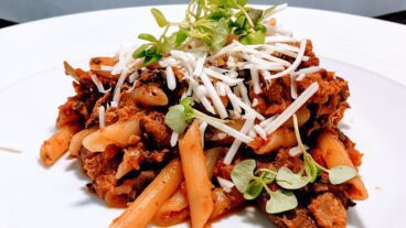 Vegan pasta bolognese on a white plate, topped with shredded cheese and microgreens.