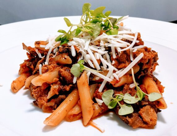 Vegan pasta bolognese on a white plate, topped with shredded cheese and microgreens.