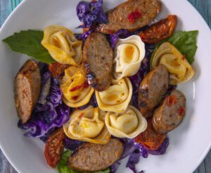 A white bowl of tortellini and sliced sausage, mixed with sundried tomatoes, purple cabbage, and basil leaves.
