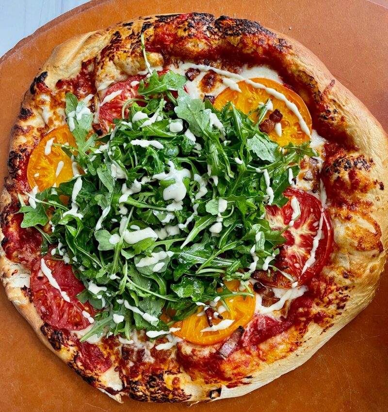 A top view of a pizza with pancetta, heirloom tomatoes, and arugula with a swirl of cream sauce.