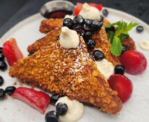 A white plate with French toast slices stacked against one another, topped with berries, dollops of cream, powdered sugar and mint.