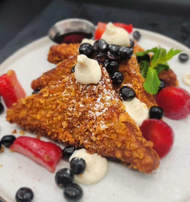 A white plate with French toast slices stacked against one another, topped with berries, dollops of cream, powdered sugar and mint.