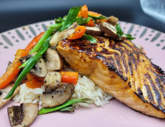 A pink plate with white rice and roasted vegetables topped with seared salmon.