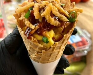A gloved hand holds a waffle cone full of mac and cheese and pulled pork, topped with BBQ sauce, green onions, and crispy onions.