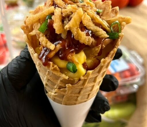 A gloved hand holds a waffle cone full of mac and cheese and pulled pork, topped with BBQ sauce, green onions, and crispy onions.