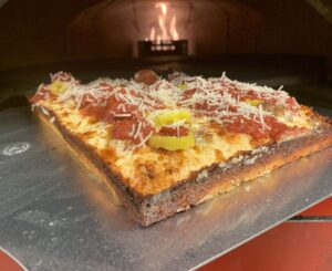 A Detroit-style pizza with pepperoni, sausage, banana peppers, and cheese staged in front of a pizza oven.
