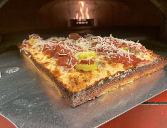 A Detroit-style pizza with pepperoni, sausage, banana peppers, and cheese staged in front of a pizza oven.