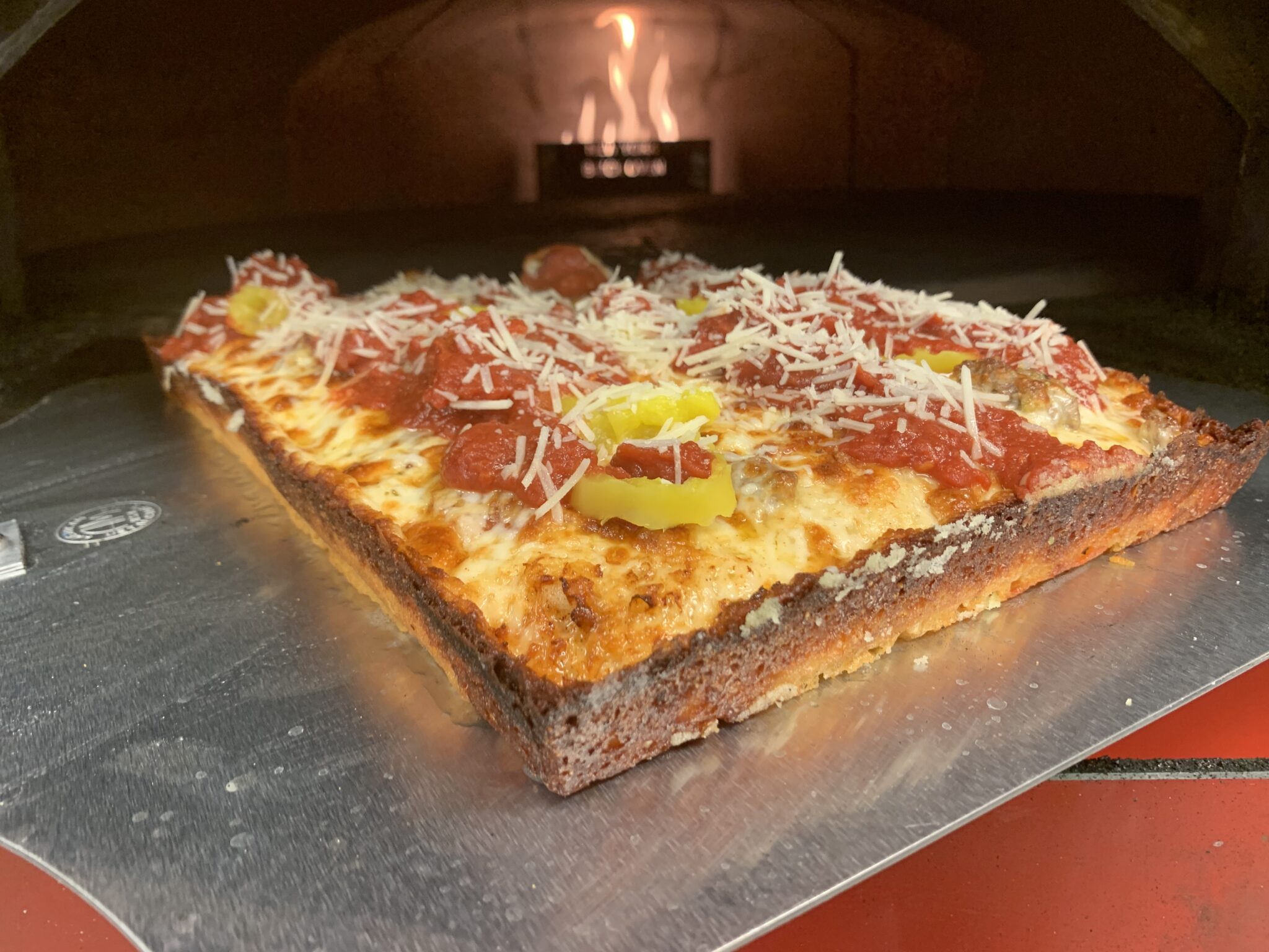 A Detroit-style pizza with pepperoni, sausage, banana peppers, and cheese staged in front of a pizza oven.
